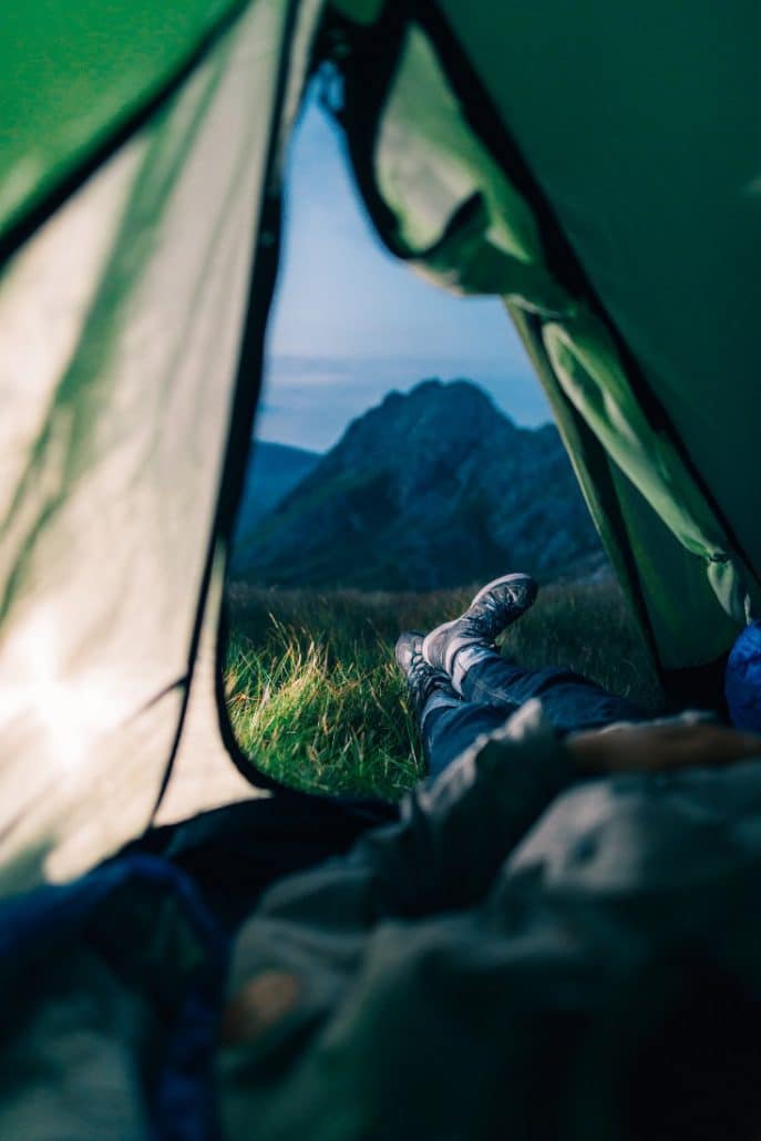View from inside a tent of crossed legs and boots facing a grassy field and dark mountain.