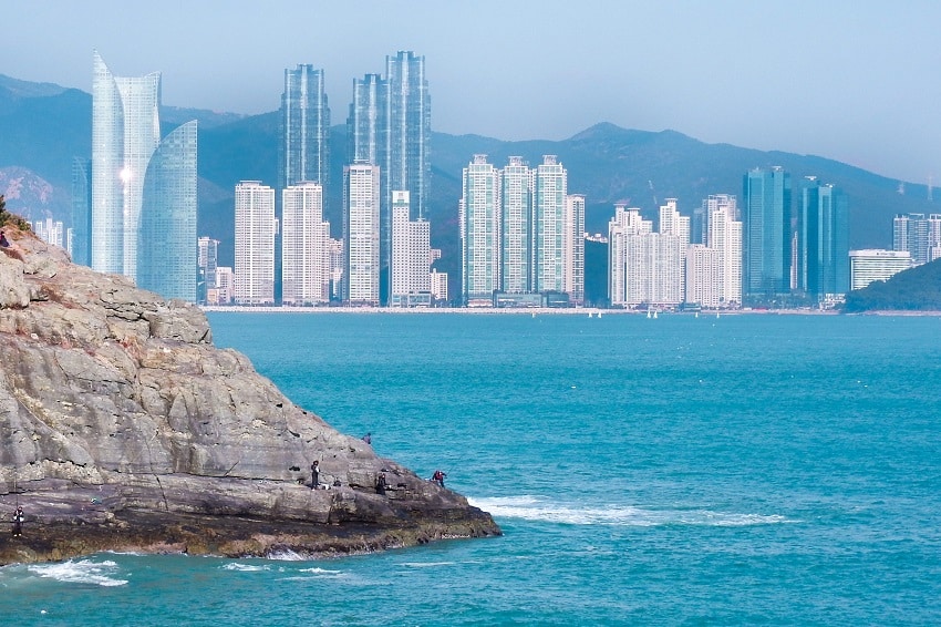 Fisherman standing on rocks fishing off Busan's East Coast with the skyscrapers of Gwangan in the background