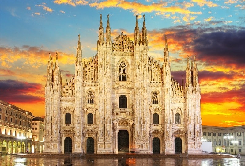 Milan Cathedral facade at sunset with an orange and purple sky behind its Gothic spires